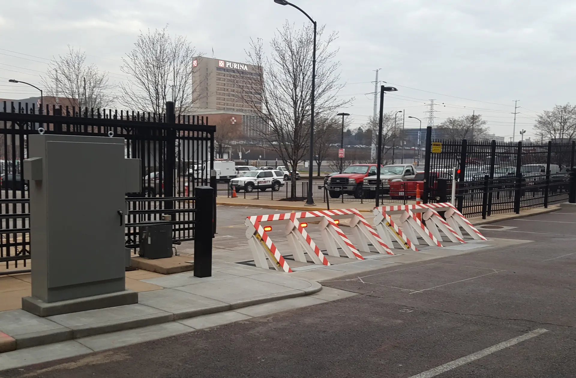 A row of white and red-striped vehicle barriers blocks an entrance near a black metal fence and a parking lot with trucks.