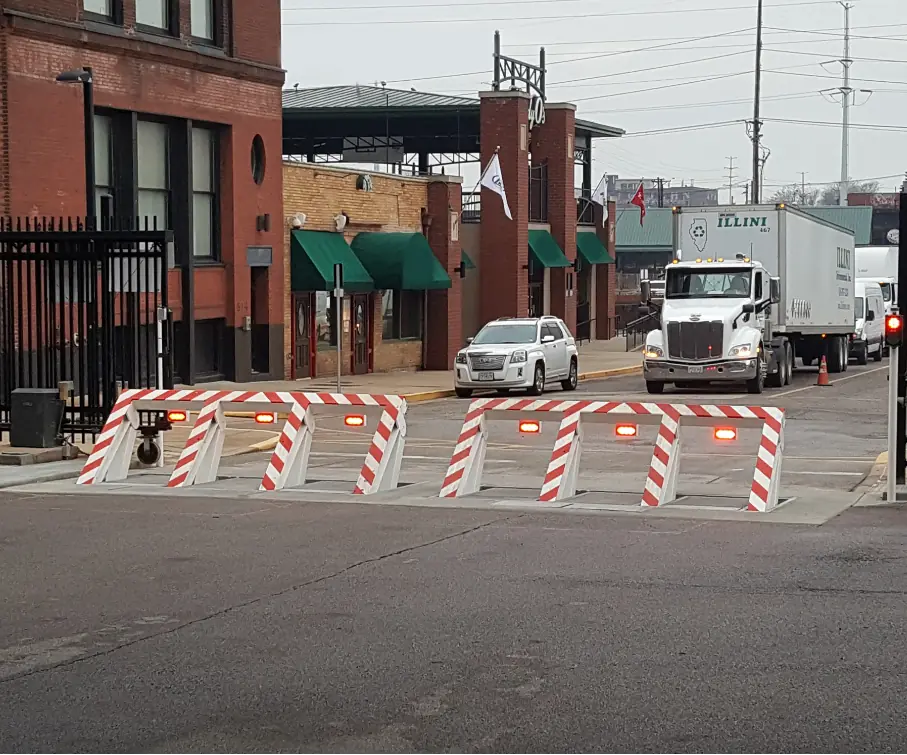 White and red-striped vehicle barriers block an entrance near a brick building and a truck waiting behind them.