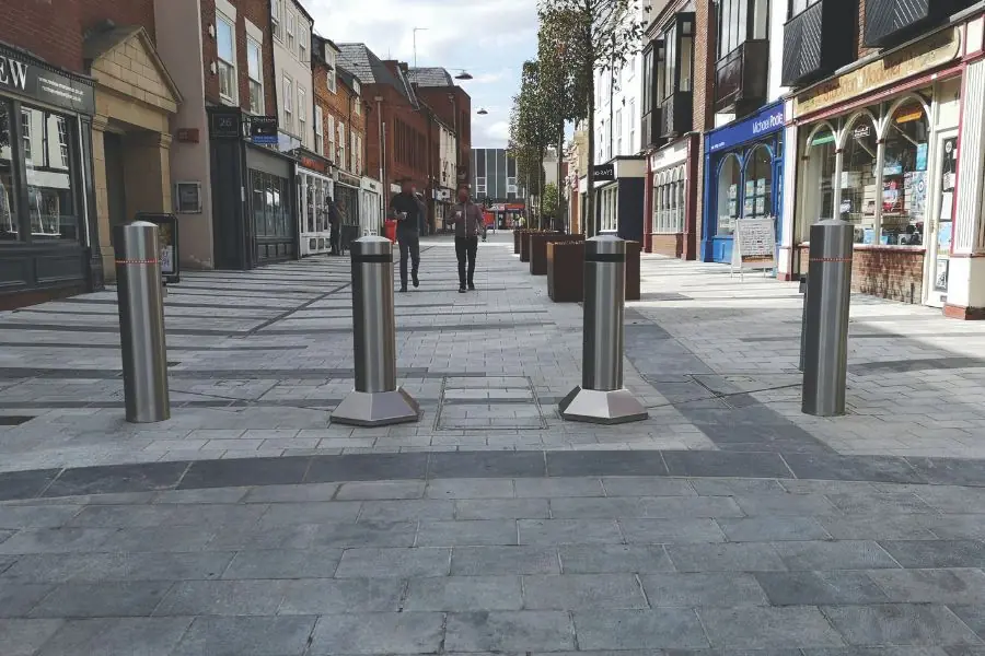 Metal bollards block vehicle access on a paved walkway with shops on both sides.