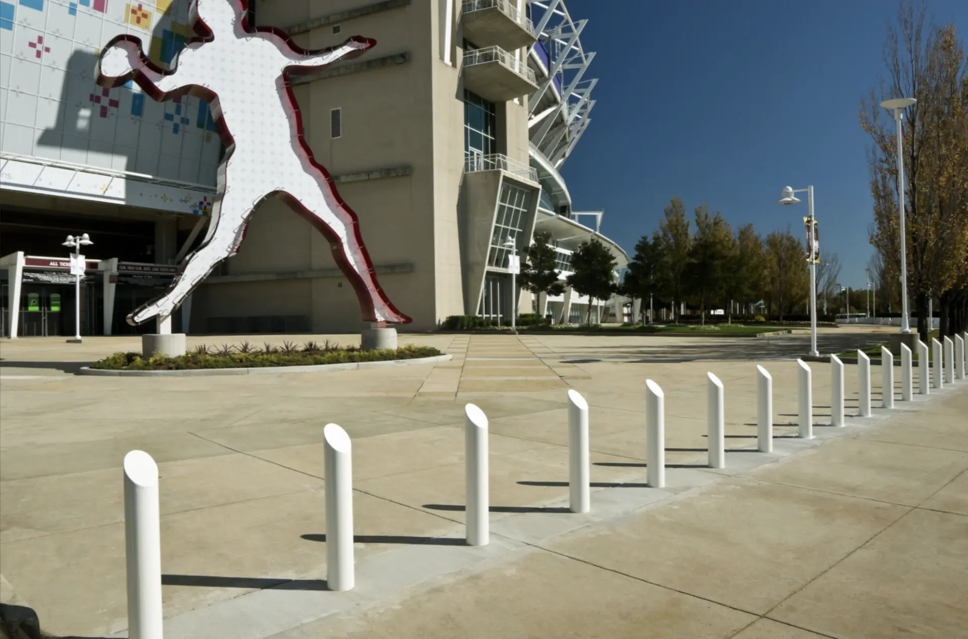 A row of white steel bollards leads toward a stadium entrance with a large figure sculpture in front.