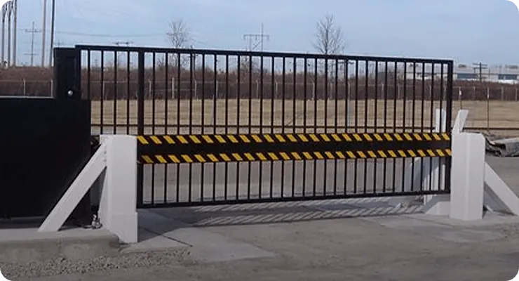 Black metal security gate with yellow and black striped barrier at a fenced perimeter.