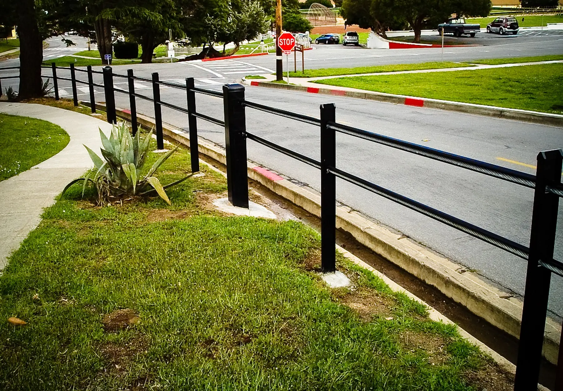 Black two-rail metal fence runs alongside a sidewalk at a street corner with grass and plants.