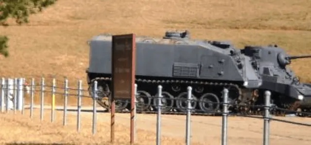 A military tank drives along a path lined with steel cable fencing in an open, grassy area.