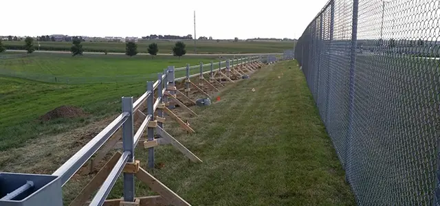 A long chain-link fence runs alongside a newly installed guardrail supported by wooden braces.