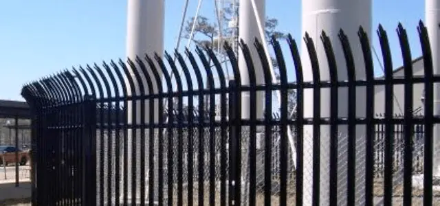 A black metal security fence with curved spikes encloses an industrial area with large pipes.