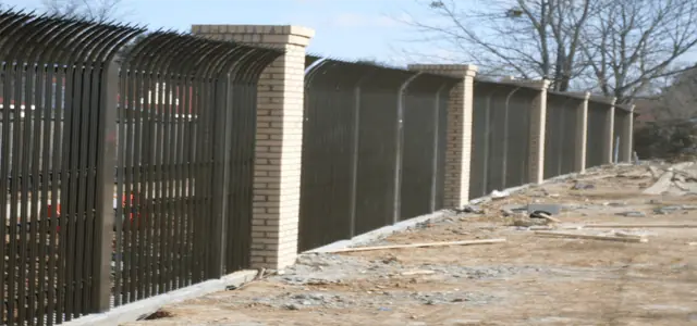 Black security fence with arched spikes and brick columns extends beside a cleared area.