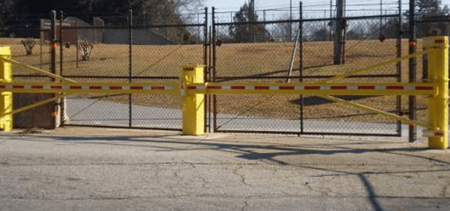 Yellow swing-arm barrier with reflective strips secures an entry point by a chain-link fence.