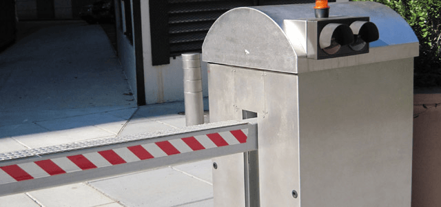 Silver security gate with red-striped arm and warning light controls access to a driveway.