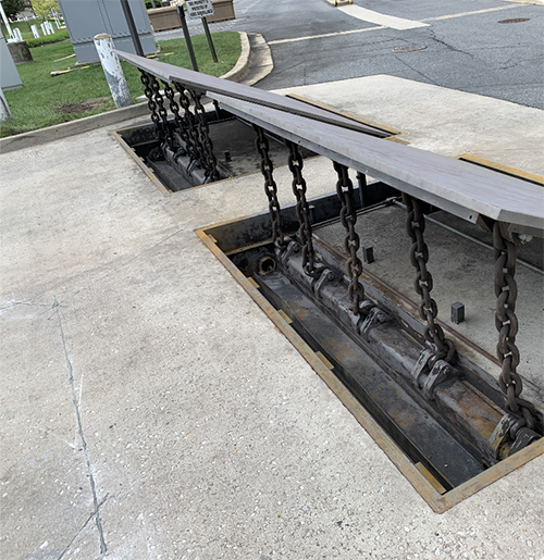 A recessed vehicle barrier with thick chains connects to a steel bar across the roadway.