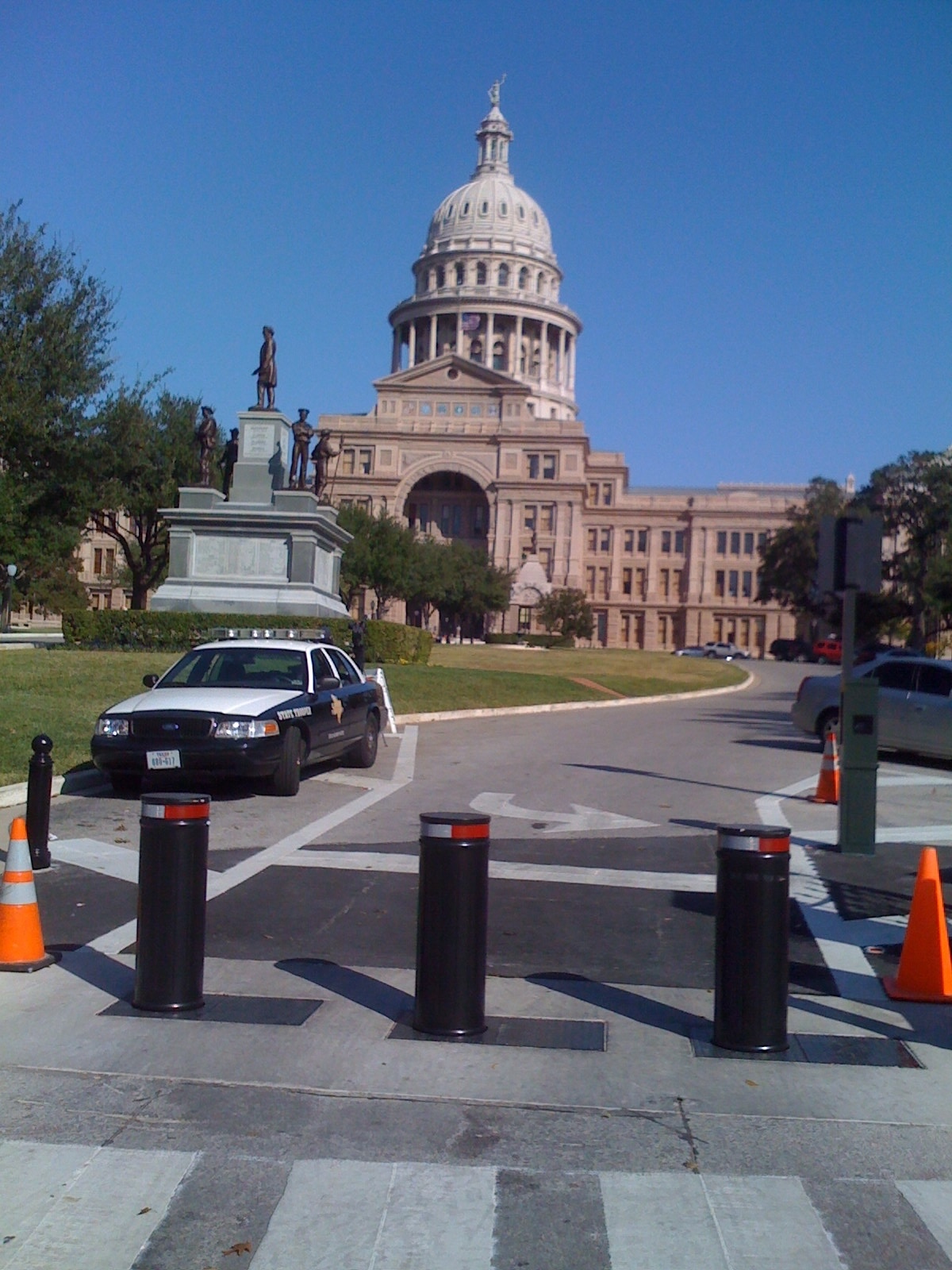 Three black retractable bollards are positioned in front of a government building, with a police car and orange cones nearby.