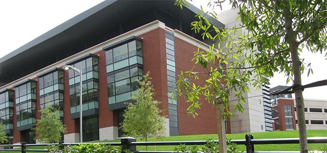 Red brick building with large windows, landscaped grounds, and black perimeter fence.