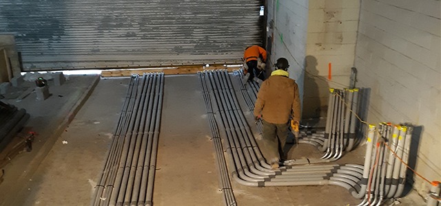 Workers install rows of conduit pipes along a concrete floor inside an industrial building.