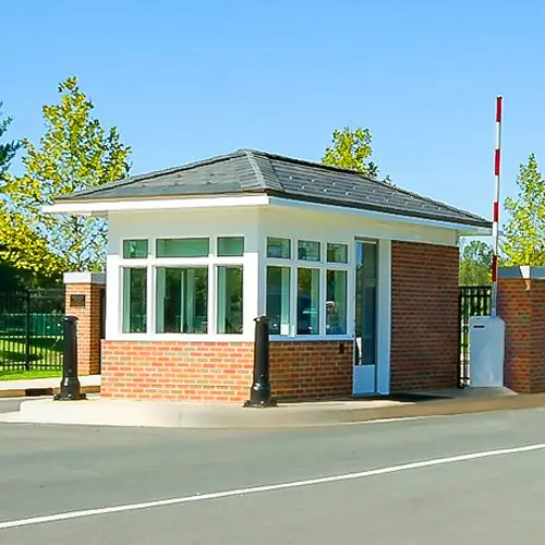 Guardhouse with a hip roof and glass walls located at the entrance of a fenced property.