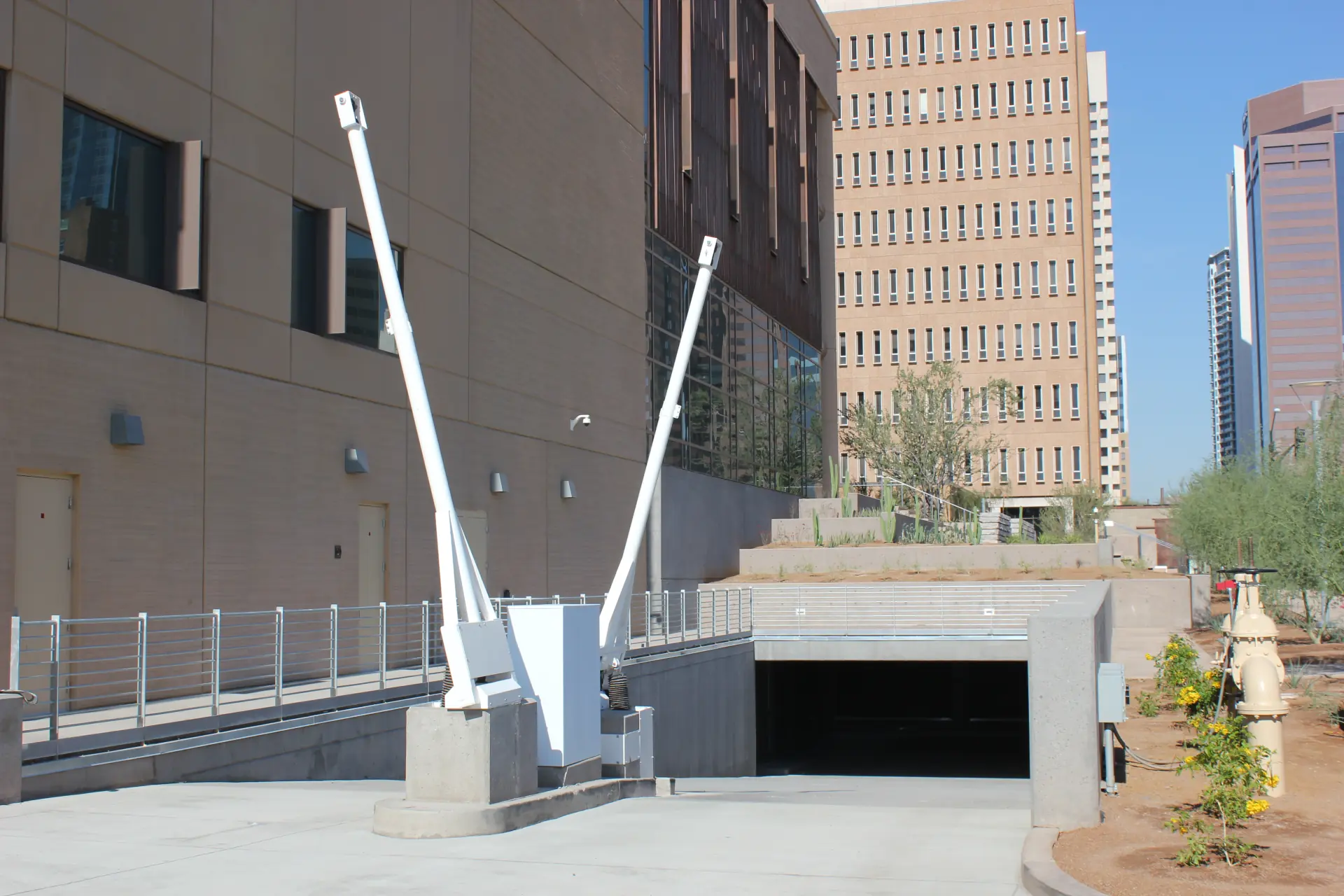 Two drop arm barriers secure a concrete ramp leading into a parking garage below urban high-rise buildings.