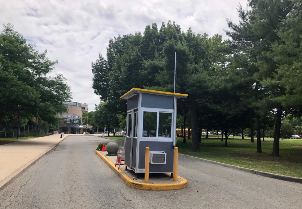 Small gray security booth with yellow roof and bollards placed on a traffic island near a tree-lined road.