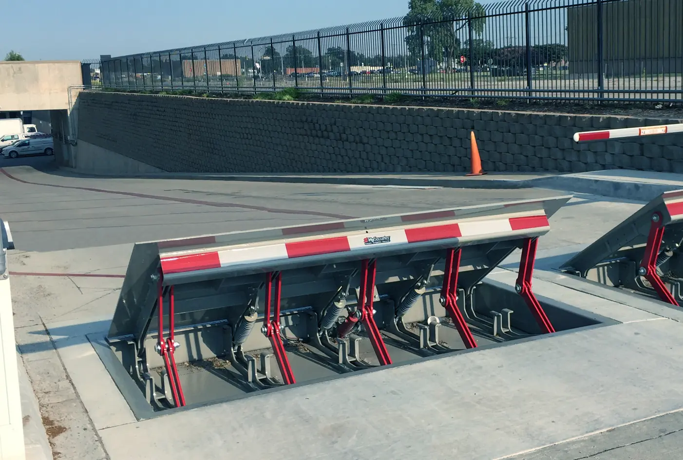 Hydraulic wedge barrier with red and white panel raised on a concrete roadway near a fenced facility.