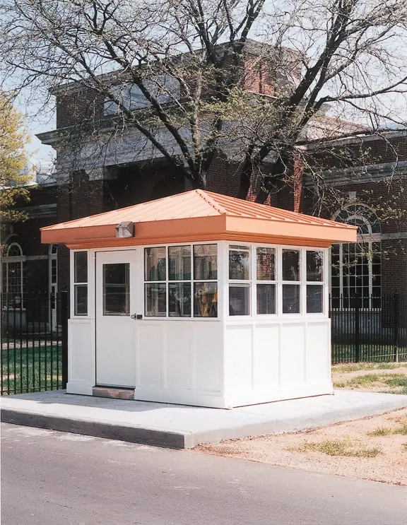 Guard booth with paneled walls and large windows positioned beside a tree-lined institutional property.