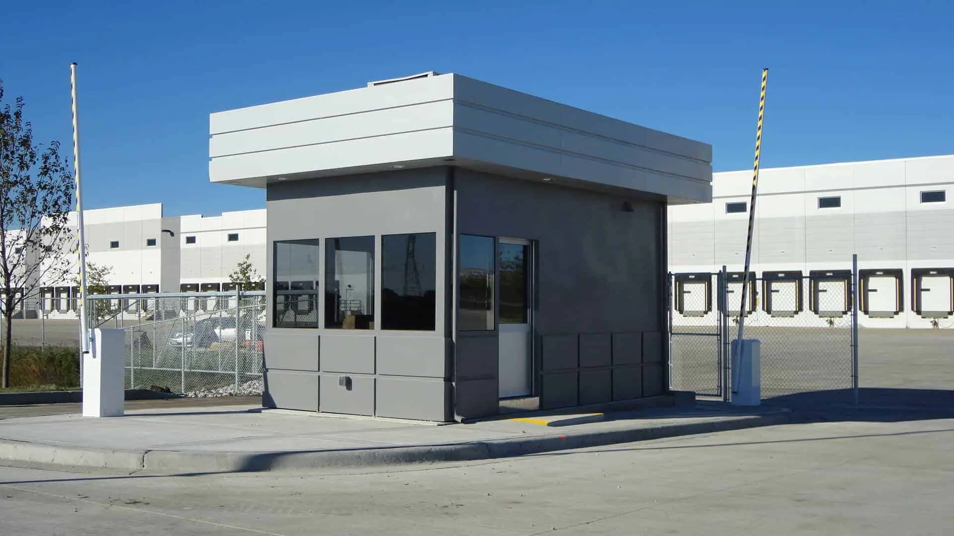 Gray security booth with flat white roof and surrounding barrier arms at a warehouse entrance.