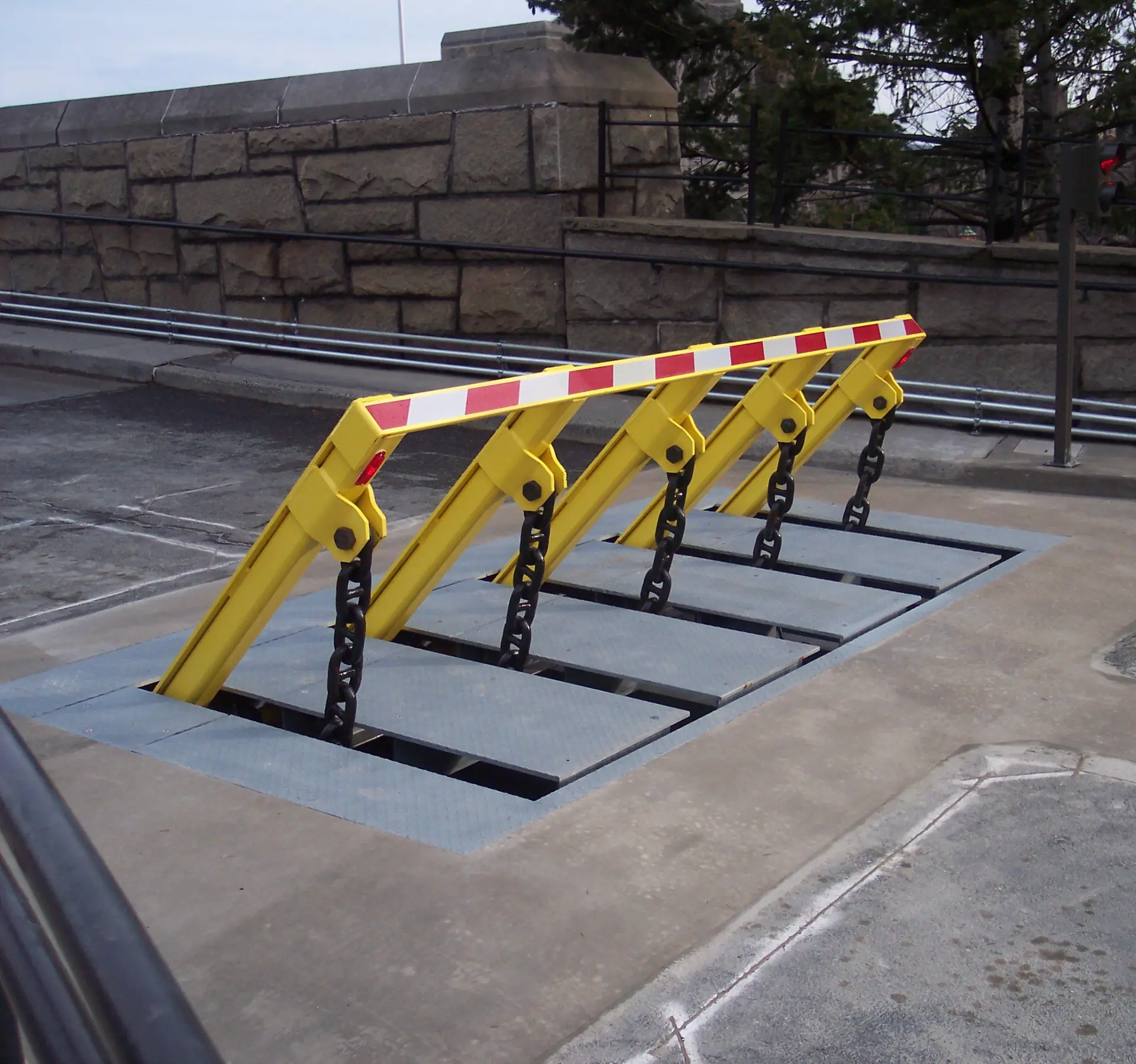 A row of yellow vehicle barriers with red-and-white reflective stripes is raised on chains in a concrete setting.