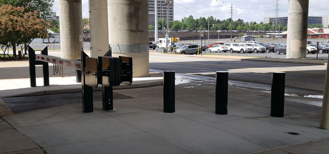 A secured roadway features bollards and a swing gate under a highway overpass.