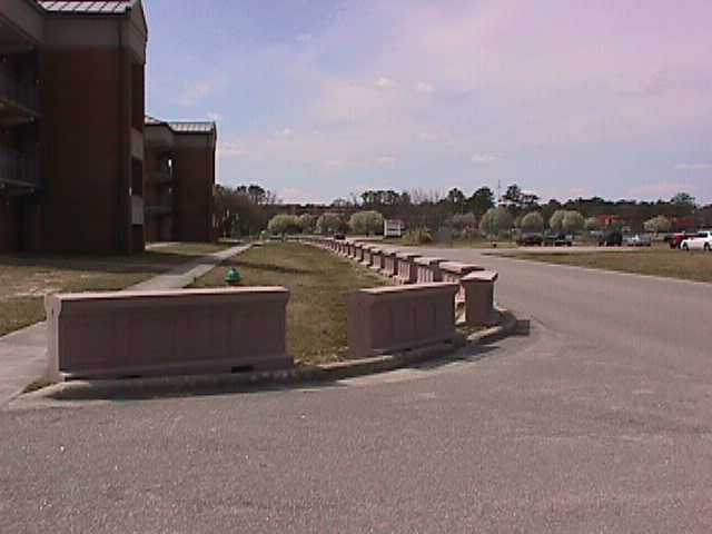 A row of rectangular concrete barriers lines the edge of a road near a residential area.