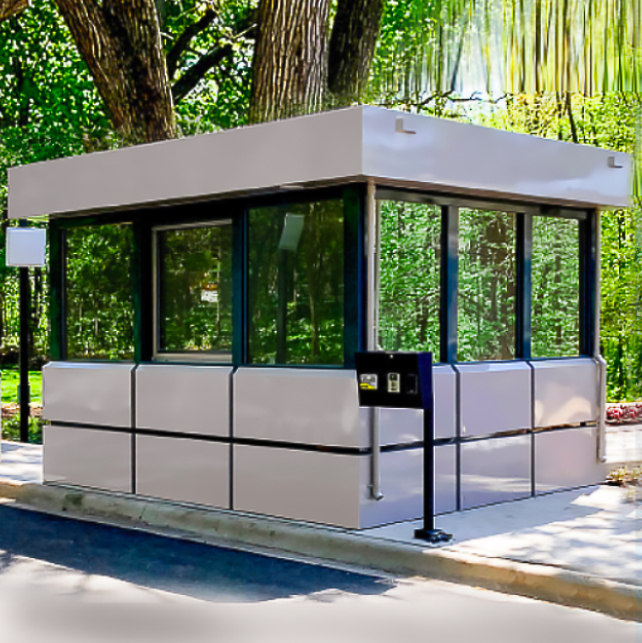 A glass guard booth with white panels sits surrounded by trees and greenery.