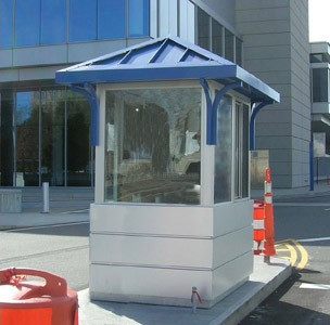 A compact white security booth with a blue metal roof sits near a modern building.