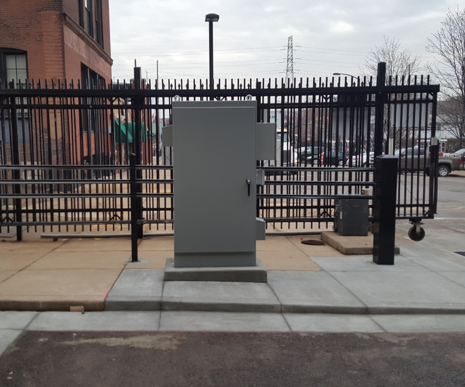 A metal security gate stands beside a gray control cabinet in an urban setting.